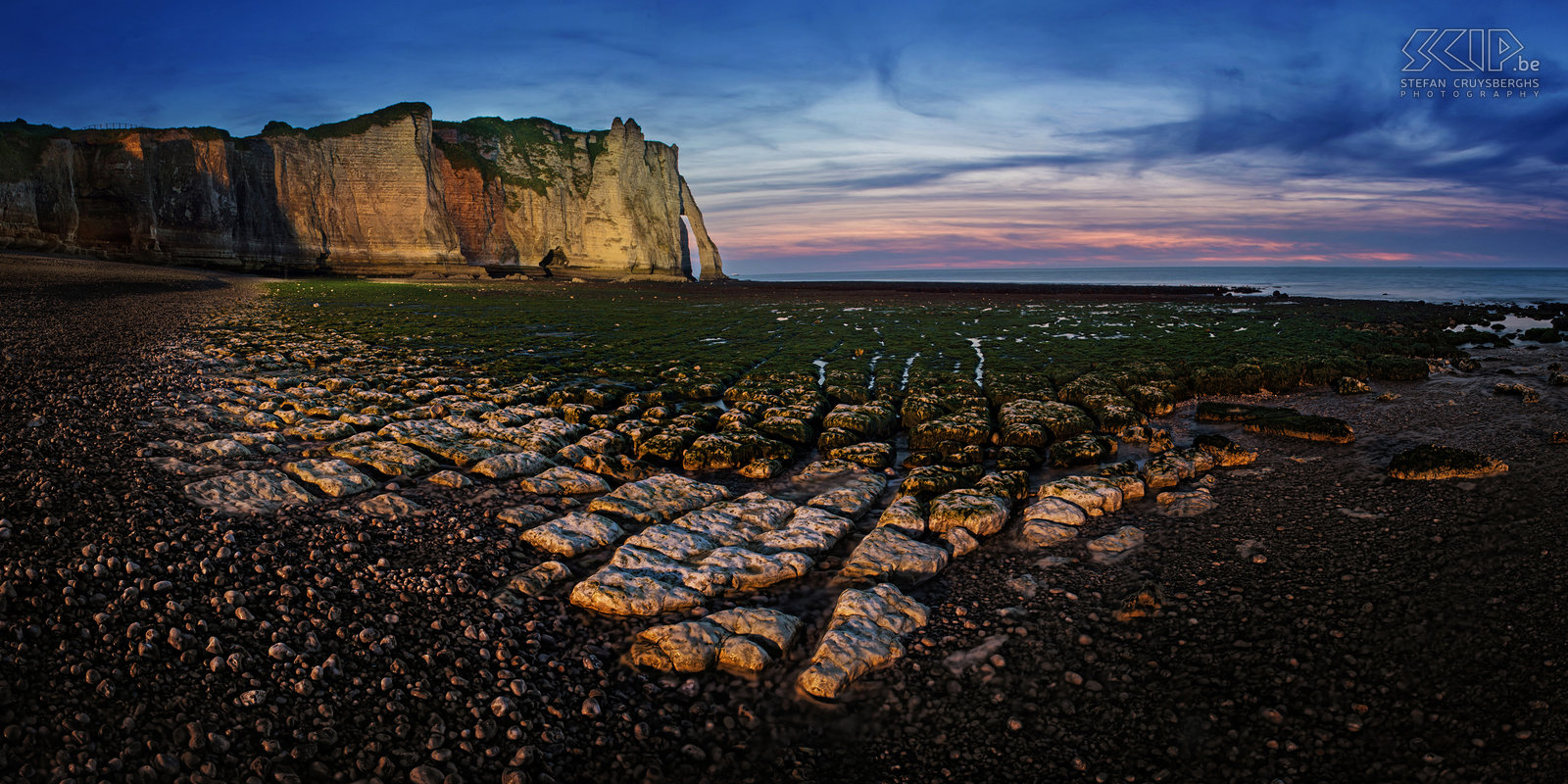 Normandy - Étretat by night We were photographing during the last light of the day and with low tide at the white cliffs of Étretat. Suddenly there was a lot of artificial light. It seems that the city has installed big spotlights to lighten up the cliffs with orange and white light during the night. This is a stiched panorama photo of the beach and cliffs. Stefan Cruysberghs